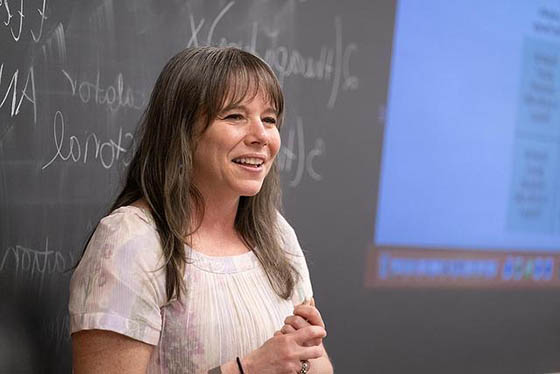 一位教授在教室前面微笑的照片, 她身后有一块黑板，在屏幕外指导学生