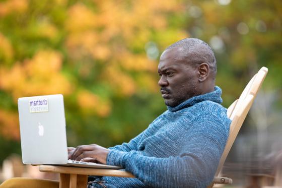 图为一名网上赌博平台的男学生在外面用笔记本电脑工作, 以秋叶为背景.