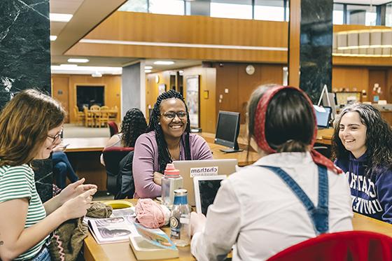 图为一群网上赌博平台的学生坐在珍妮·金·梅隆图书馆的学习桌旁，微笑着一起学习. 