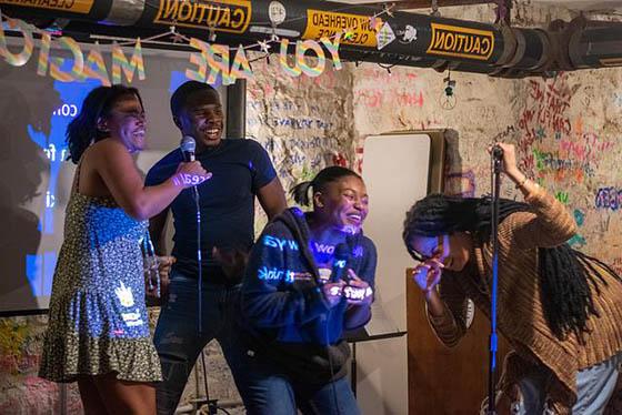 在BIPOC卡拉ok之夜，四名黑人学生拿着麦克风在舞台上笑着唱歌的照片. 在他们后面有一个标志，说你是神奇的.